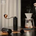 Molino de café manual acero inoxidable junto a un kit de café de goteo con hielo y vapor en una mesa de madera.