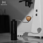 Molino de café manual acero inoxidable de color negro con manivela de madera bajo una luz artística con sombras.