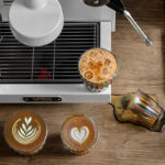 Vista superior de cuatro vasos de vidrio MUVNA sobre barra de madera, incluyendo el vaso multicolor para café 320 ml con diferentes bebidas y arte latte.